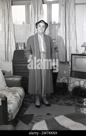 1950er, historisch, eine ältere Dame in einem langen Wollmantel, Hut, Handschuhe und Handtasche in der Hand, steht in einem Vorderzimmer, ein kleiner Fernseher aus der Epoche in der Ecke, möglicherweise eines vom britischen Hersteller Ferranti, England, Großbritannien. Stockfoto