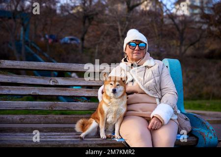 Ziemlich reife Frau mit Sonnenbrille und Shiba Inu Hund, der auf einer Holzbank sitzt Stockfoto