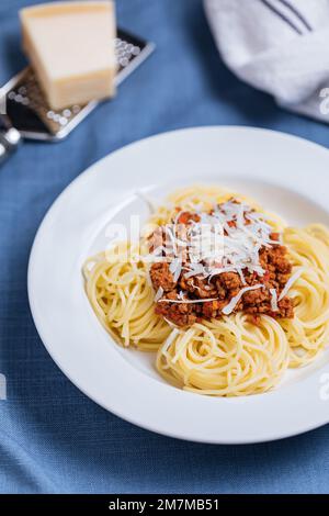 Bolognese-Spaghetti mit Parmesankäse auf weißem Teller. Einfacher blauer Hintergrund und Dekoration. Traditionelles italienisches Gericht, mediterrane Küche Stockfoto