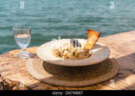 Meeresfrüchte-Pasta-Spaghetti und ein Glas Wasser stehen auf einem Holztisch im Café-Restaurant mit Landschaft Meer im Hintergrund. Leer kopieren Stockfoto