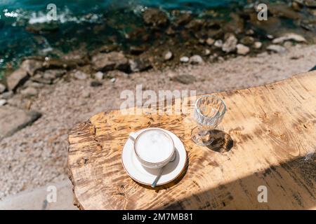 Cappuccino Latte Kaffeetasse und ein Glas Wasser stehen auf einem Holztisch im Café draußen mit Landschaft Meer und Berge im Hintergrund. Kopieren Stockfoto