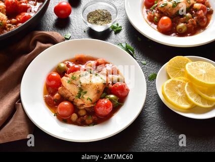 Kabeljaueintopf mit Kichererbsen, Kirschtomaten und Oliven auf dem Teller auf dunklem Steingrund. Stockfoto