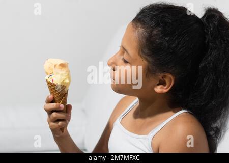 Ein kleines braungehäutetes Latina-Mädchen, das eine Eiskrem-Tüte in der Hand hält und sie sich ansieht, bevor sie sie isst Stockfoto