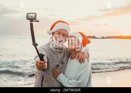 Porträt von zwei süßen alten Menschen, die Spaß haben und gemeinsam am Strand an weihnachtstagen am Strand mit Weihnachtshüten genießen. Ich schaue und halte eine Kamera, um Videos von Ferien aufzunehmen Stockfoto