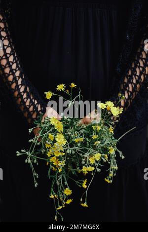 Schwarz gekleidete Frau mit gelben Wildblumen hinter ihrem Rücken Stockfoto