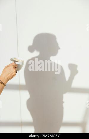 Weibliche Hand hält Kaffeetasse, während ihr Schatten an der Wand zu sehen ist Stockfoto
