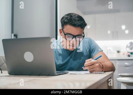 Ein junger Teenager, der abends zu Hause auf dem Tisch lernt und arbeitet, mit einem lächelnden Laptop und viel Spaß bei den Hausaufgaben. Stockfoto