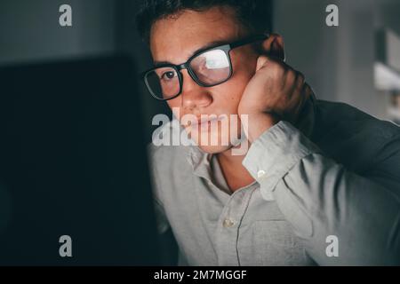 Nahaufnahme und Porträt eines jungen Teenagers, der abends zu Hause auf dem Tisch lernt und arbeitet, indem er einen lächelnden Laptop benutzt und Spaß bei den Hausaufgaben hat. Licht des Bildschirms auf dem Gesicht. Stockfoto