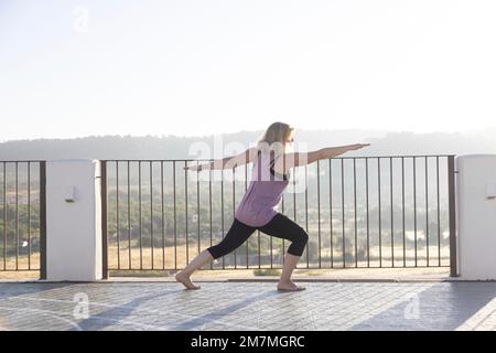 Blonde Frau, die bei Sonnenaufgang Yoga auf einer Terrasse in Spanien praktiziert Stockfoto