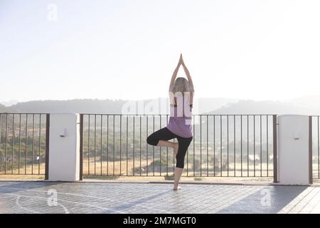 Blonde Frau, die bei Sonnenaufgang Yoga auf einer Terrasse in Spanien praktiziert Stockfoto