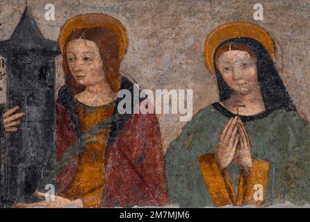 St. Barbara (links) und St. Margarete von Cortona. Fresko gemalt 1512 an der Westfront einer ehemaligen Kapelle Cappella di San Grato in Aosta, Aostatal, Italien. Die Heilige Barbara hält ihr heiliges Attribut, einen Turm mit drei Fenstern und einem konischen Dach, Symbol der Gefangenschaft ihres Vaters und der Heiligen Dreifaltigkeit. Stockfoto