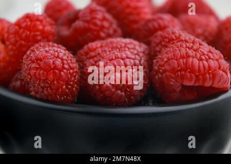 Nahaufnahme frischer Himbeeren in dunkler Schüssel, verschwommener weißer Hintergrund Stockfoto