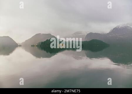 Einsame Häuser auf einer Insel in Norwegen, Landschaft mit Reflexion im Wasser, Nebel und Wolken, typische Fjordlandschaft mit kleinen Inseln, Abgeschiedenheit von der Außenwelt, zentrale Perspektive Stockfoto