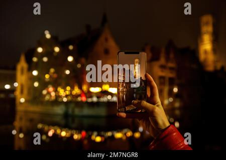 Fotografieren Sie die belgische Altstadt von Brügge, die bei Nacht beleuchtet ist. Tourist mit Smartphone Stockfoto