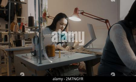 Weibliche Schneiderin in Brille sitzt an der Nähmaschine und arbeitet an maßgeschneiderter Kleidung. Arbeiten Sie Kollegen am Tisch mit einem Laptop im Hintergrund näher. Atelier oder Nähwerkstatt. Modebegriff. Stockfoto