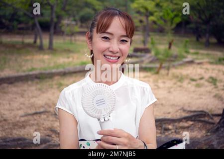 Eine reife Japanerin draußen in einem Park an einem heißen Tag mit einem kleinen elektrischen Ventilator. Stockfoto