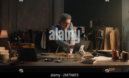 Ein erwachsener Mann entwirft mit Tablet und Laptop ein Kleidungsdesign für die neue Bekleidungskollektion. Tailor in Brille schaut in die Kamera, steht neben dem Tisch mit Schneidewerkzeugen. Konzept von Mode und Handwerk. Stockfoto