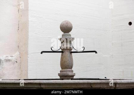 Ein alter Steinbrunnen steht vor einer grob verputzten Hauswand. Die Säule und die Wasserspeier sind dekoriert. Stockfoto