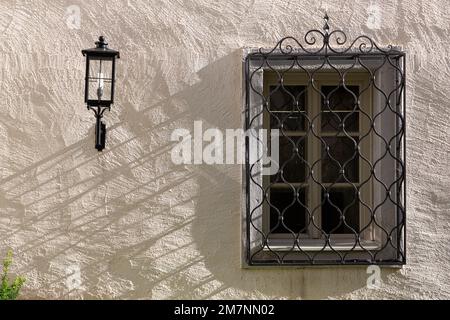 Das Fenster in einer grob verputzten Hauswand ist mit einem schmiedeeisernen Gitter befestigt. Eine antike Lampe ist an der Wand montiert. Stockfoto