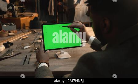 Afroamerikanischer Schneider sitzt am Tisch in einem luxuriösen Atelier, surft im Internet oder zeichnet Kleidungsdesign auf einem Tablet-Computer mit grünem Bildschirm und Eingabestift. Chromakey. Nahaufnahme. Stockfoto