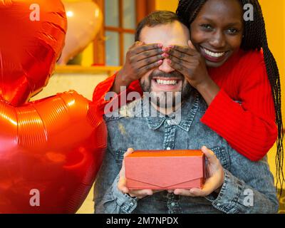 Porträt eines gemischtrassigen Paares mit einem Mann, der überrascht ist, während ihm seine Freundin oder Frau am Valentinstag ein Geschenk schenkt Stockfoto