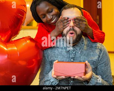 Porträt eines gemischtrassigen Paares mit einem Mann, der überrascht ist, während ihm seine Freundin oder Frau am Valentinstag ein Geschenk schenkt Stockfoto