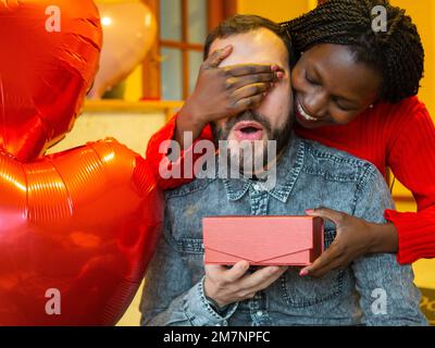 Porträt eines gemischtrassigen Paares mit einem Mann, der überrascht ist, während ihm seine Freundin oder Frau am Valentinstag ein Geschenk schenkt Stockfoto