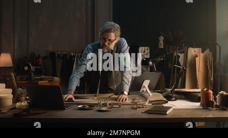 Ein erwachsener Mann entwirft mit Tablet und Laptop ein Kleidungsdesign für die neue Bekleidungskollektion. Tailor in Brille schaut in die Kamera, steht neben dem Tisch mit Schneidewerkzeugen. Konzept von Mode und Handwerk. Stockfoto