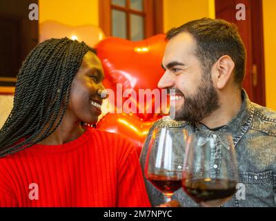 Porträt eines gemischtrassigen Paares, das sich mit Gläsern Rotwein zum Valentinstag zum Lachen ansieht Stockfoto