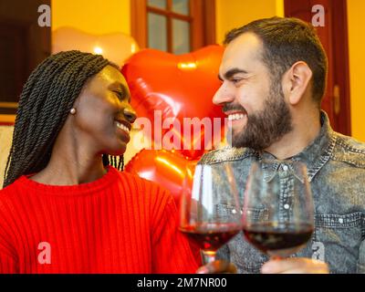 Porträt eines gemischtrassigen Paares, das sich mit Gläsern Rotwein zum Valentinstag zum Lachen ansieht Stockfoto