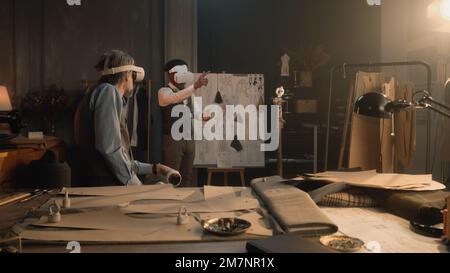 Multiethnische Schneider, die ein VR-Headset mit schnurlosen Controllern tragen. Sie arbeiten in einem Luxusatelier oder in einem Spezialstudio und erstellen Business Suit in virtueller Realität. Technologiekonzept in der Wirtschaft Stockfoto