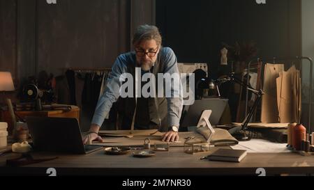 Ein erwachsener Mann entwirft mit Tablet und Laptop ein Kleidungsdesign für die neue Bekleidungskollektion. Tailor in Brille schaut in die Kamera, steht neben dem Tisch mit Schneidewerkzeugen. Konzept von Mode und Handwerk. Stockfoto