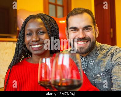 Porträt eines gemischtrassigen Paares, das in die Kamera schaut und mit Gläsern Rotwein lacht und den Valentinstag genießt Stockfoto