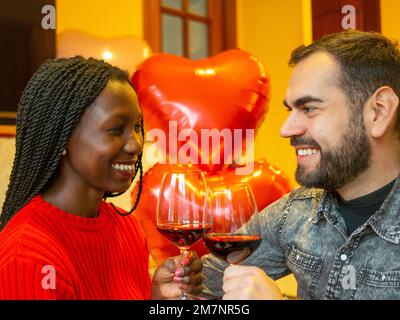 Porträt eines gemischtrassigen Paares, das sich mit Gläsern Rotwein zum Valentinstag zum Lachen ansieht Stockfoto