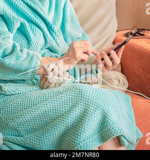 Nahaufnahme von Frauenhänden mit Smartphone beim Stricken mit Nadeln. Konzept des handgemachten Hobbys, Technologie Stockfoto