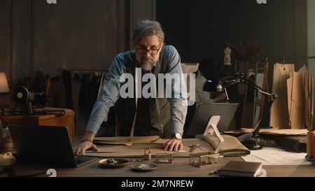 Ein erwachsener Mann entwirft mit Tablet und Laptop ein Kleidungsdesign für die neue Bekleidungskollektion. Tailor in Brille schaut in die Kamera, steht neben dem Tisch mit Schneidewerkzeugen. Konzept von Mode und Handwerk. Stockfoto