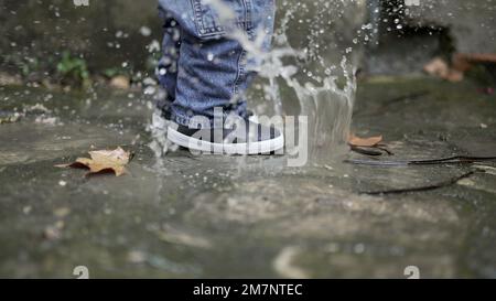 Ein Kind, das in eine Wasserpfütze springt. Ein Kleinkind, das Stiefel trägt, spritzt in die Pfütze Stockfoto