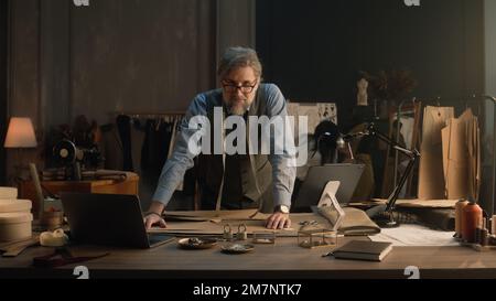 Ein erwachsener Mann entwirft mit Tablet und Laptop ein Kleidungsdesign für die neue Bekleidungskollektion. Tailor in Brille schaut in die Kamera, steht neben dem Tisch mit Schneidewerkzeugen. Konzept von Mode und Handwerk. Stockfoto