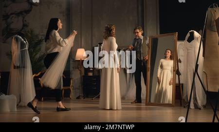 Filmaufnahme eines weiblichen Schneiders, der der Frau im Hochzeitskleid vor dem Spiegel den Schleier aufsetzt. Braut im Designer-Atelier oder im Hochzeitssalon ein passendes Hochzeitskleid. Modebegriff und Couturier. Stockfoto