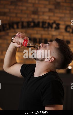 RIVNE, UKRAINE JUNI 26 2020: Junger bärtiger Mann trinkt Coca Cola aus der Flasche im Friseurladen auf Backsteinmauer Hintergrund. Stockfoto