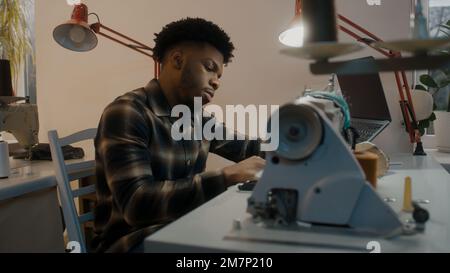 Afroamerikanischer Schneider oder Näherin sitzt an der Nähmaschine mit Laptop am Tisch. Männermode-Designer arbeitet an einem modernen Hochzeitsanzug. Atelier oder Nähwerkstatt. Modebegriff. Seitenschießen. Stockfoto