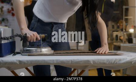 Frau bügelt Textilien in Atelier-Werkstatt. Ihre Kollegen arbeiten im Hintergrund: Der Mann arbeitet mit maßgeschneiderten Anzügen, die Schneiderin arbeitet an der Nähmaschine. Konzept von Mode und Handwerk. Stockfoto