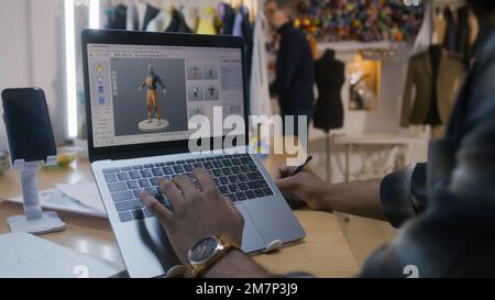 Afroamerikanischer Modedesigner kreiert Kleidung in 3D-Modelliersoftware auf einem Laptop mit digitalem Tablet und Eingabestift. Er arbeitet in der Atelier-Werkstatt. Mode- und Technologiekonzept in der Wirtschaft. Stockfoto