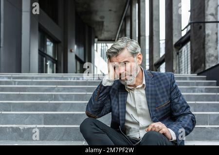 Grauhaariger Geschäftsmann, der traurig auf der Treppe vor dem Bürogebäude saß, deprimierter Chef im Business-Anzug, verärgert und enttäuscht von den Ergebnissen. Stockfoto