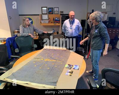 Susan Anderson (rechts) bietet einige Einblicke in die Flagge des Bundesstaates Indiana, die sie als Artefakt an das Hampton Roads Naval Museum spendet, an den Museumserzieher Zach Smyers (Mitte) und die stellvertretende Direktorin Toni Deetz-Rock (links). Die Flagge gehörte Andersons Vater, dem verstorbenen Captain Walter "Jack" Deal, USN, der während des Vietnamkriegs der befehlshabende Offizier des River Assault Squadron 15 war. Damals war es üblich, dass Bootskapitäne während des Vietnamkriegs die Staatsflagge ihres Heimatstaates auf ihren Flussangriffsbooten führten. Stockfoto