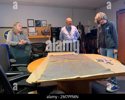 Susan Anderson (rechts) bietet einige Einblicke in die Flagge des Bundesstaates Indiana, die sie als Artefakt an das Hampton Roads Naval Museum spendet, an den Museumserzieher Zach Smyers (Mitte) und die stellvertretende Direktorin Toni Deetz-Rock (links). Die Flagge gehörte Andersons Vater, dem verstorbenen Captain Walter "Jack" Deal, USN, der während des Vietnamkriegs der befehlshabende Offizier des River Assault Squadron 15 war. Damals war es üblich, dass Bootskapitäne während des Vietnamkriegs die Staatsflagge ihres Heimatstaates auf ihren Flussangriffsbooten führten. Stockfoto