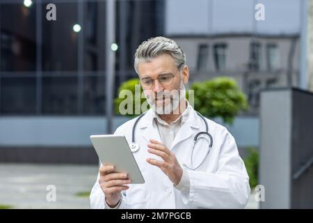 Seriöser reifer Arzt im medizinischen Kleid außerhalb der modernen Klinik mit Tablet-Computer für Videoanrufe, Mann mit Stethoskop konsultiert Patienten online. Stockfoto