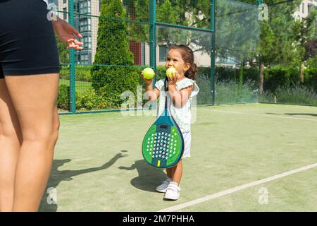 Ein kleines Mädchen, das seiner Mutter Tennisbälle auf einem Padel-Tennisplatz gibt Stockfoto