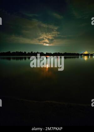 Farbfotografie der abendlichen Natur am Fluss. Aufsteigender Vollmond über den Bäumen. Wunderschöne Reflexion des Mondwegs im Wasser. Stockfoto