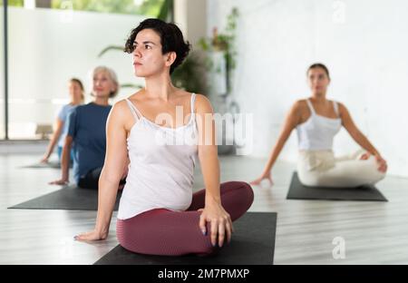 Junge, lateinische Sportlerin, die sich in Ardha Matsyendrasana während des Gruppen-Yoga-Trainings verdreht Stockfoto